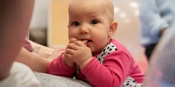 Urocze, 4-miesięczne dziecko w różowej bluzeczce z serduszkami wkłada rączki do buzi, zaciekawione światem.